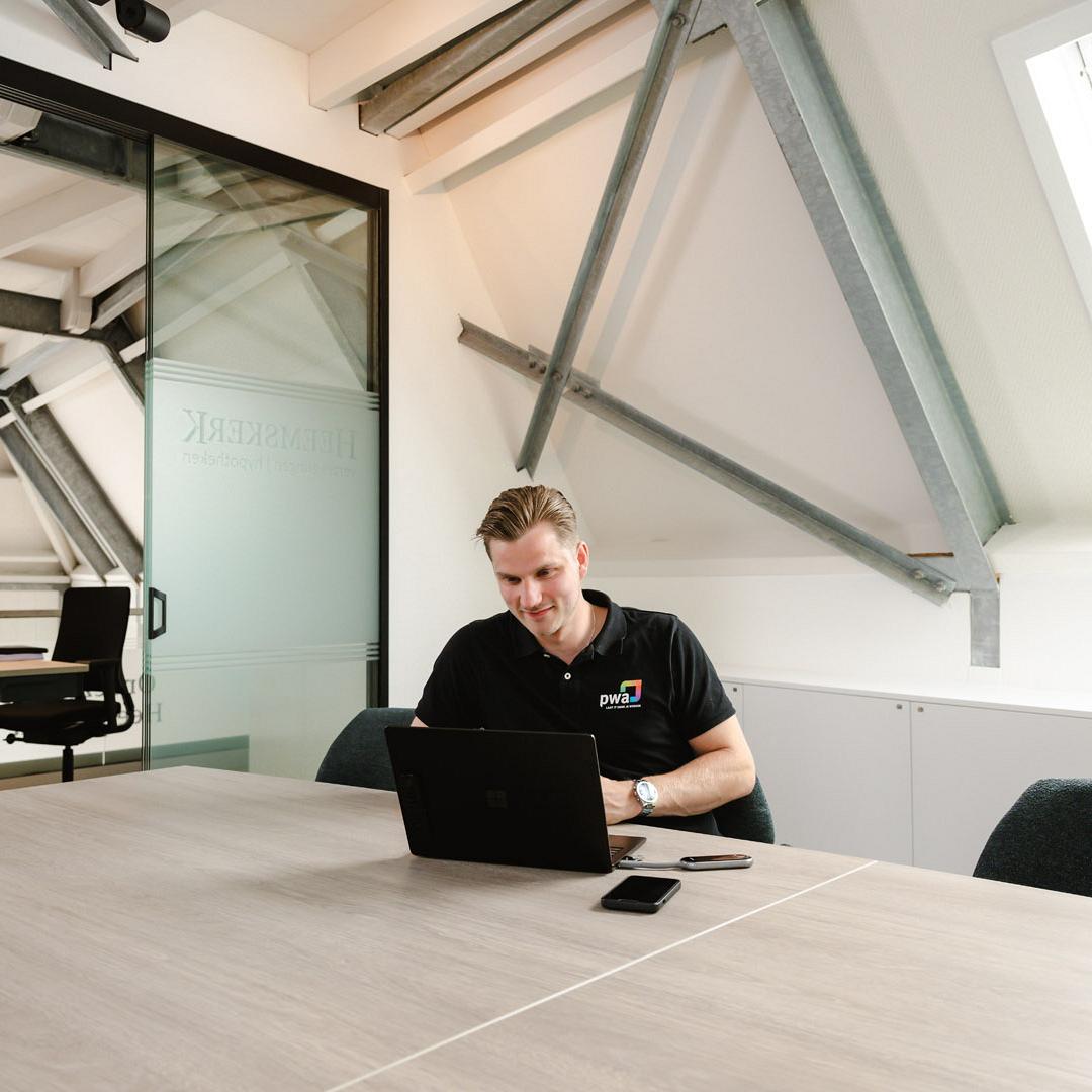 Man in PWA-shirt werkt geconcentreerd op een laptop aan een vergadertafel in een modern kantoor met schuine balken en glazen wanden.