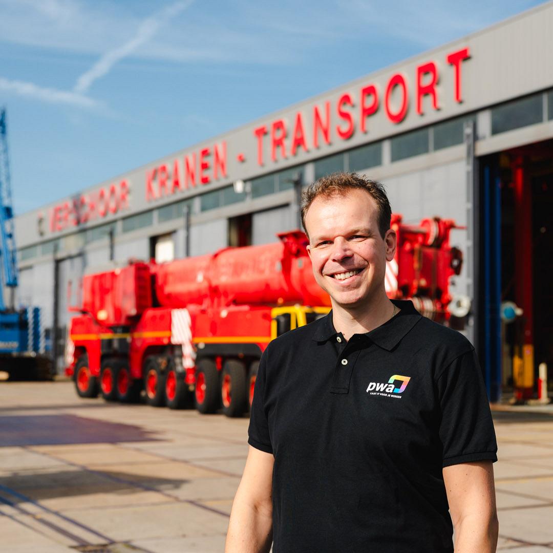 Lachende man in PWA-shirt staat voor een werkplaats van een kraan- en transportbedrijf met opvallende rode en blauwe voertuigen op de achtergrond.