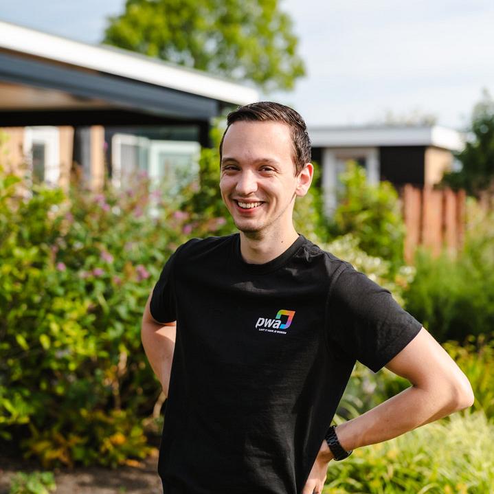 Lachende man in PWA-shirt poseert buiten voor een tuin met bloemen en struiken op een zonnige dag.