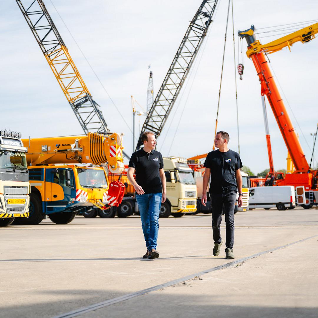 Twee mannen in bedrijfskleding lopen over een kraanterrein met imposante hijskranen en vrachtwagens op de achtergrond.