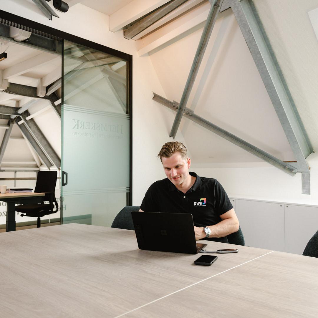 Man in PWA-shirt werkt geconcentreerd op een laptop aan een vergadertafel in een modern kantoor met schuine balken en glazen wanden.