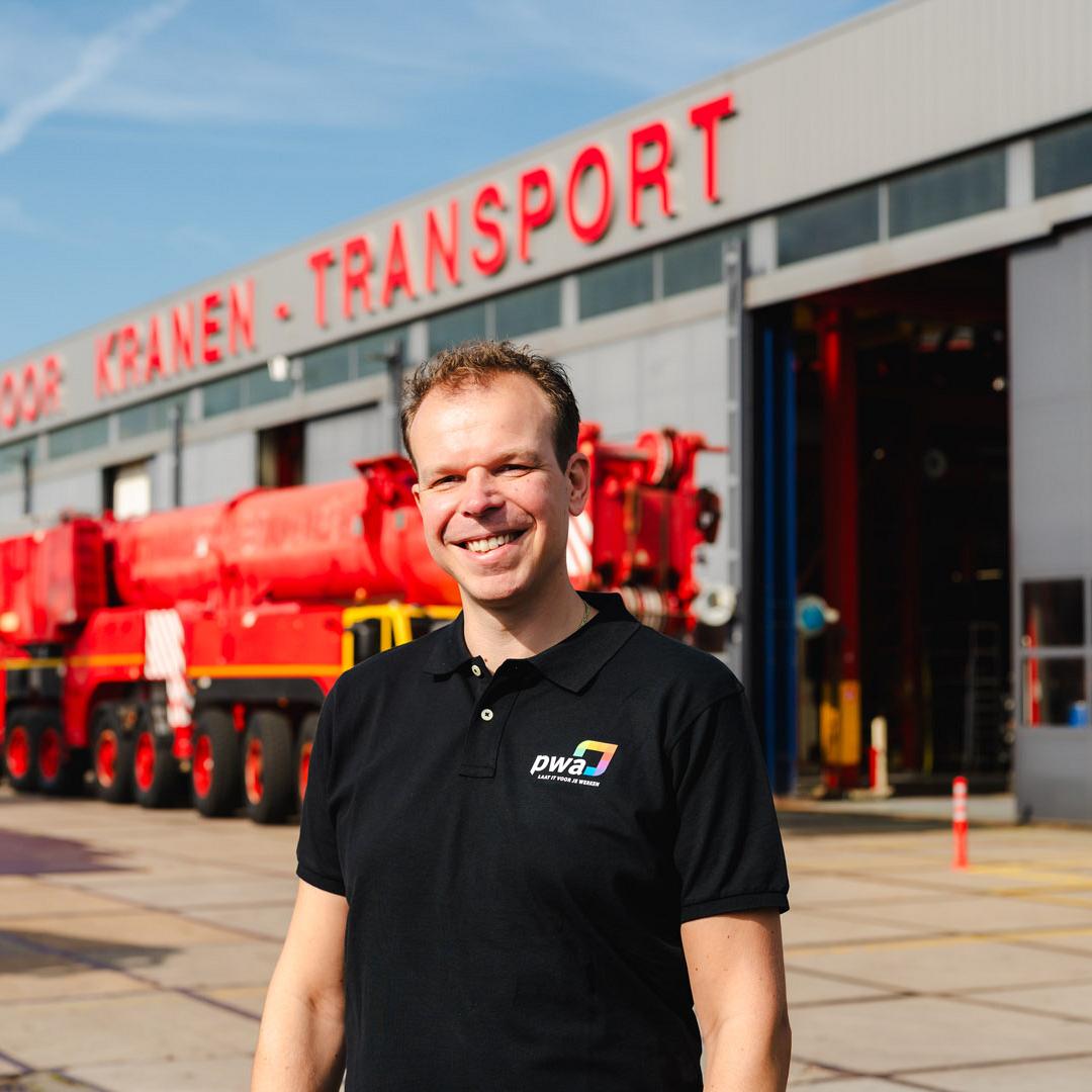 Lachende man in PWA-shirt staat voor een werkplaats van een kraan- en transportbedrijf met opvallende rode en blauwe voertuigen op de achtergrond.