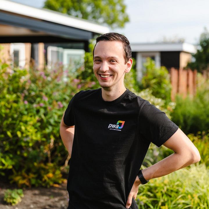 Lachende man in PWA-shirt poseert buiten voor een tuin met bloemen en struiken op een zonnige dag.