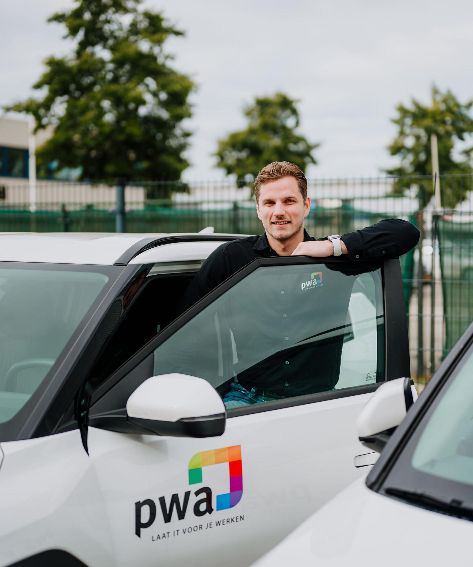 Man in PWA-bedrijfskleding leunt tegen de open deur van een witte auto met PWA-logo op de zijkant, op een bedrijventerrein met bomen en hekwerk op de achtergrond.
