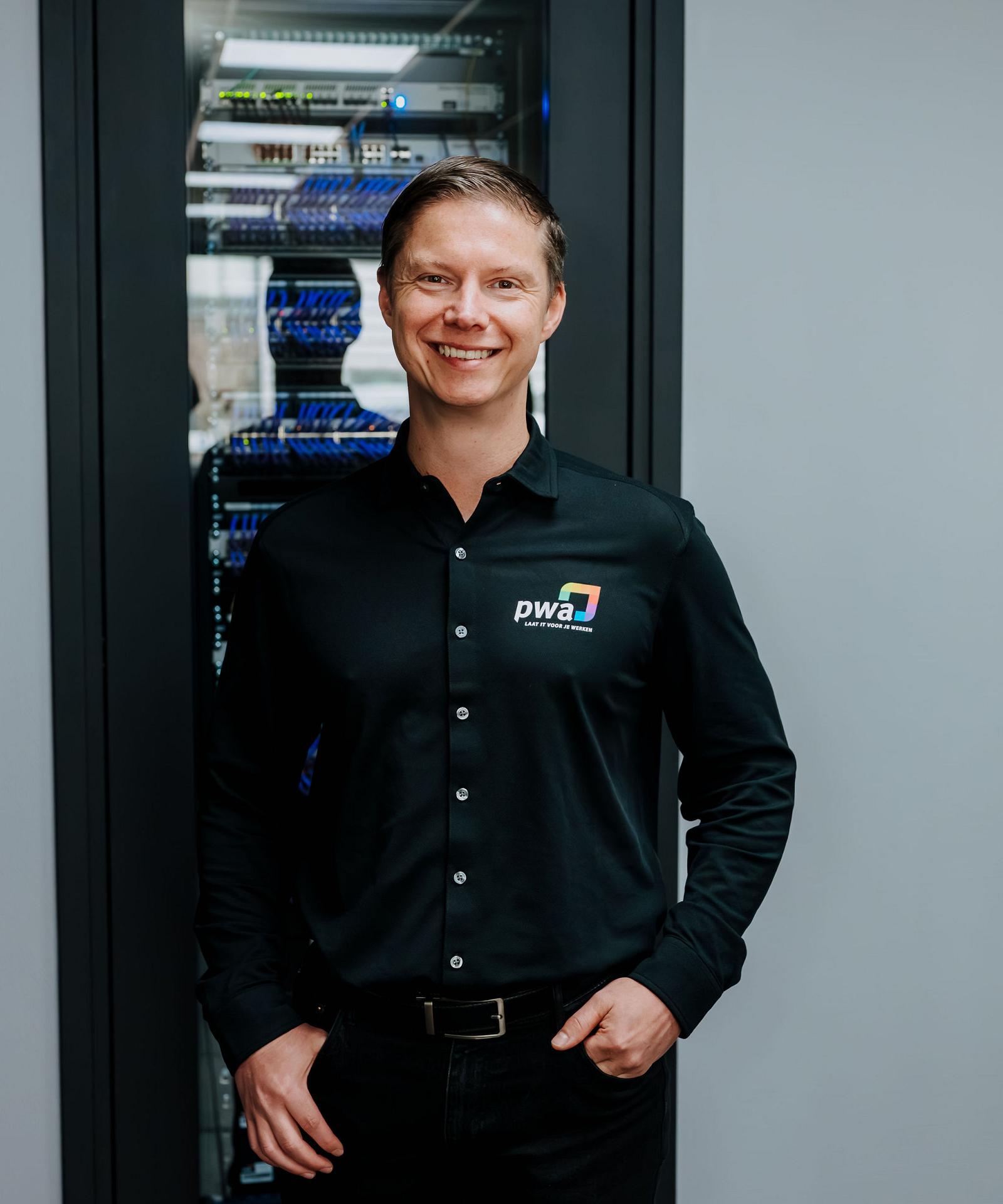 Man in PWA-bedrijfskleding staat glimlachend voor een serverkast met blauwe netwerkkabels in een moderne IT-ruimte.