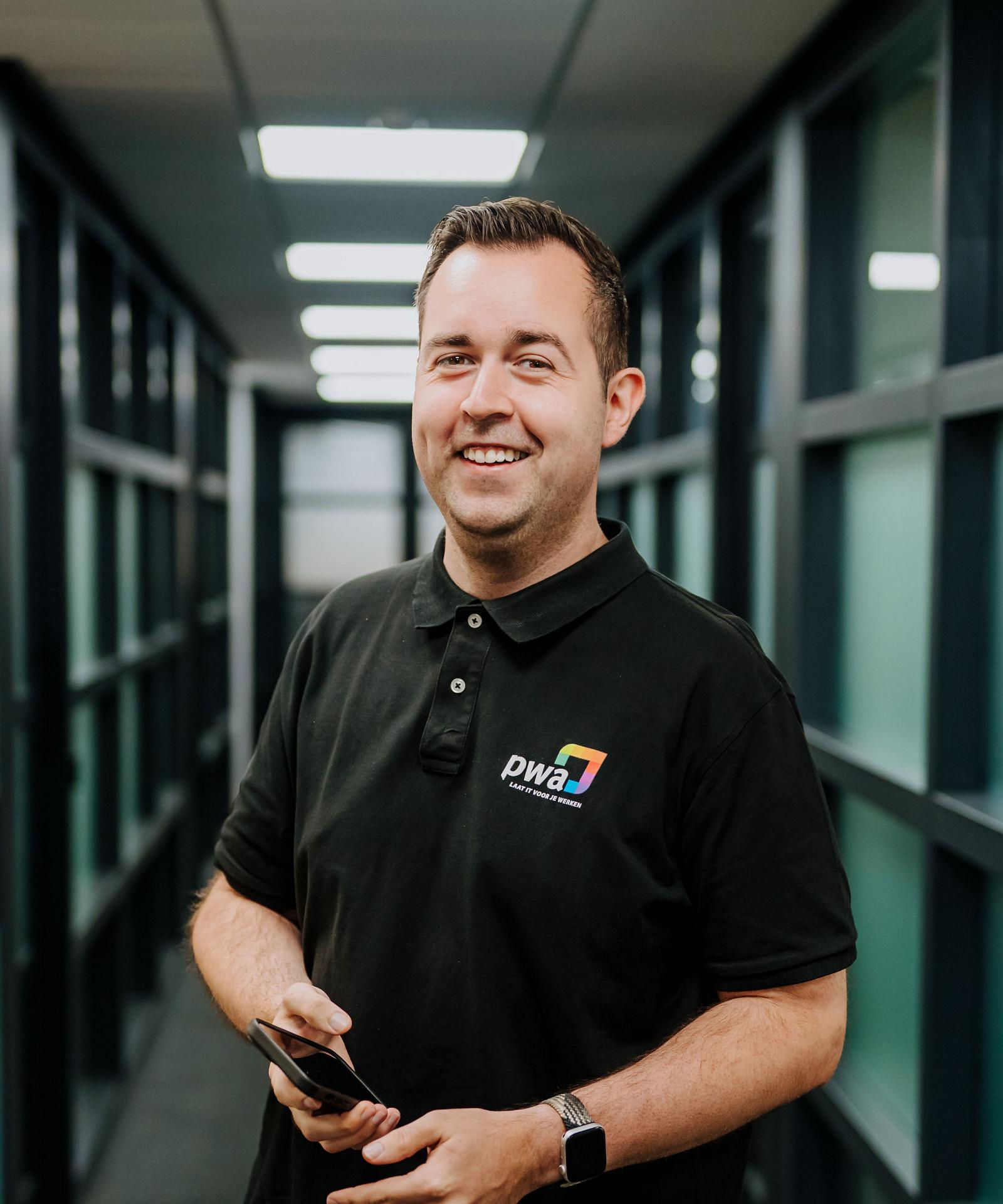 Man in PWA-bedrijfskleding staat lachend in een kantoorhal met glazen wanden en houdt een smartphone in zijn handen.