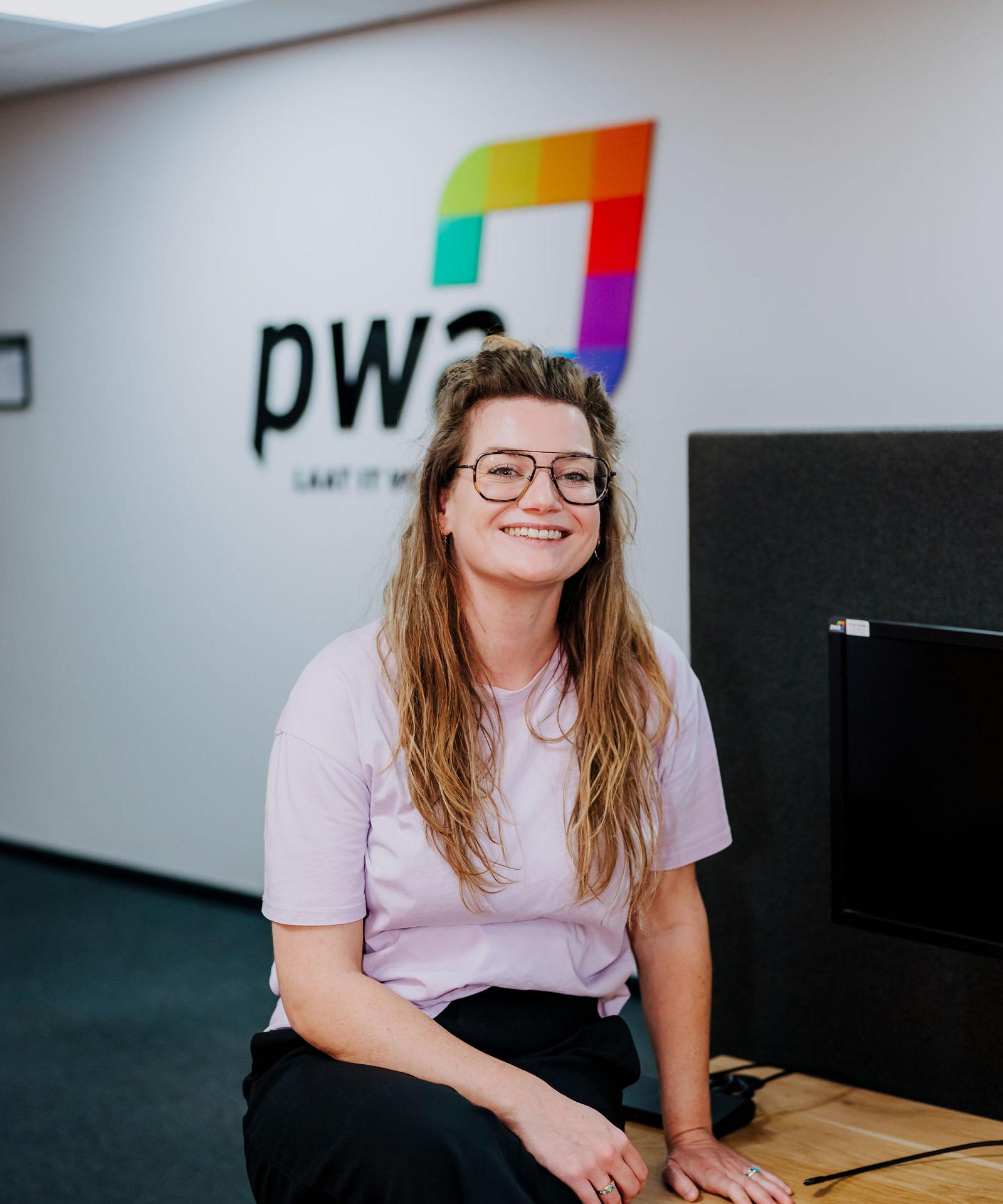 Vrouw met bril en lang haar glimlacht terwijl ze op een bureau zit in het PWA-kantoor, met het PWA-logo zichtbaar op de achtergrond.