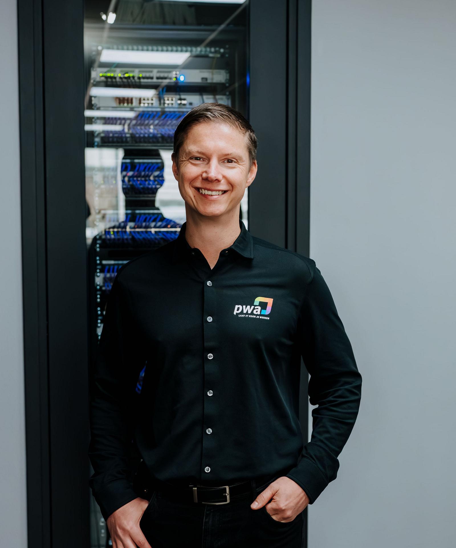 Man in PWA-bedrijfskleding staat glimlachend voor een serverkast met blauwe netwerkkabels in een moderne IT-ruimte.