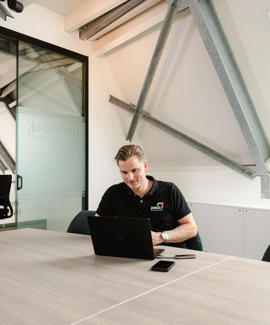 Man in PWA-shirt werkt geconcentreerd op een laptop aan een vergadertafel in een modern kantoor met schuine balken en glazen wanden.