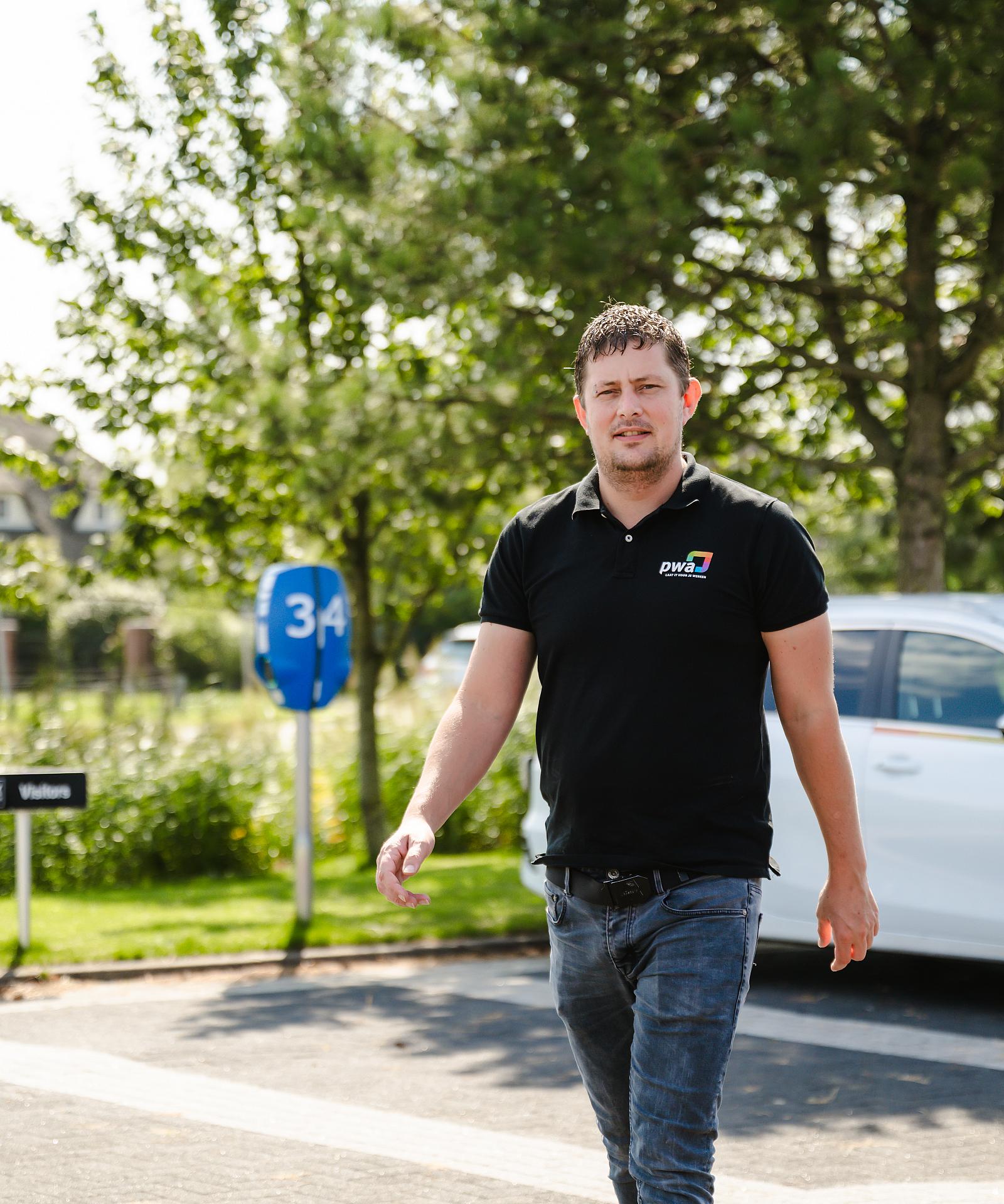 IT-specialist van PWA arriveert op locatie, wandelend vanaf een PWA-bedrijfsauto op een zonnige dag."