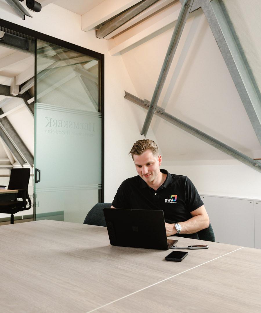 Man in PWA-shirt werkt geconcentreerd op een laptop aan een vergadertafel in een modern kantoor met schuine balken en glazen wanden.