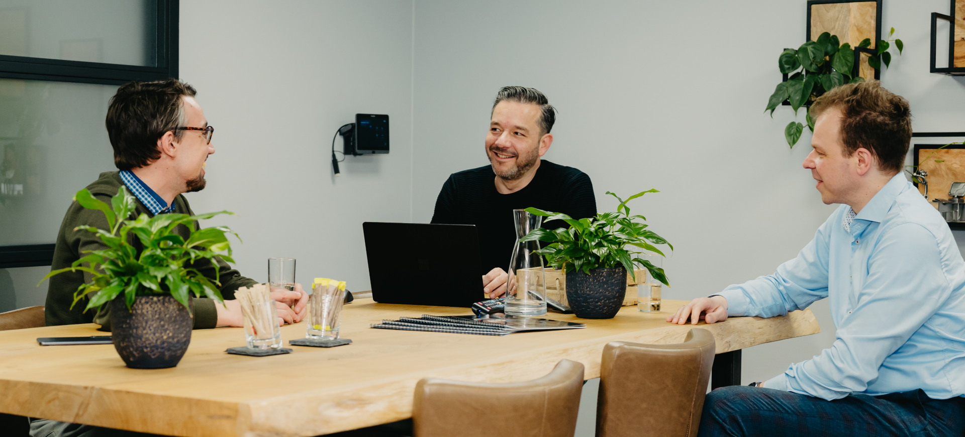 Drie mannen zitten aan een houten vergadertafel met een laptop, notitieboeken en glazen water, en hebben een informeel overleg in een moderne kantoorruimte.