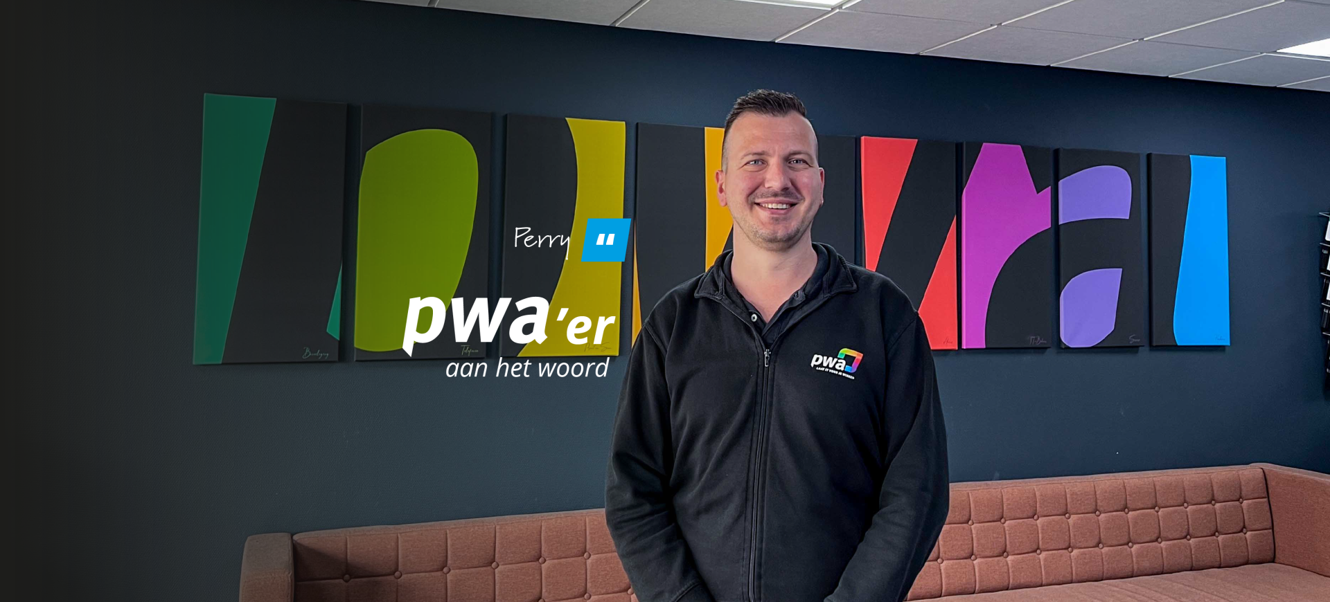 Perry, PWA’er aan het woord, lachend voor een kleurrijke wand in het PWA-kantoor.