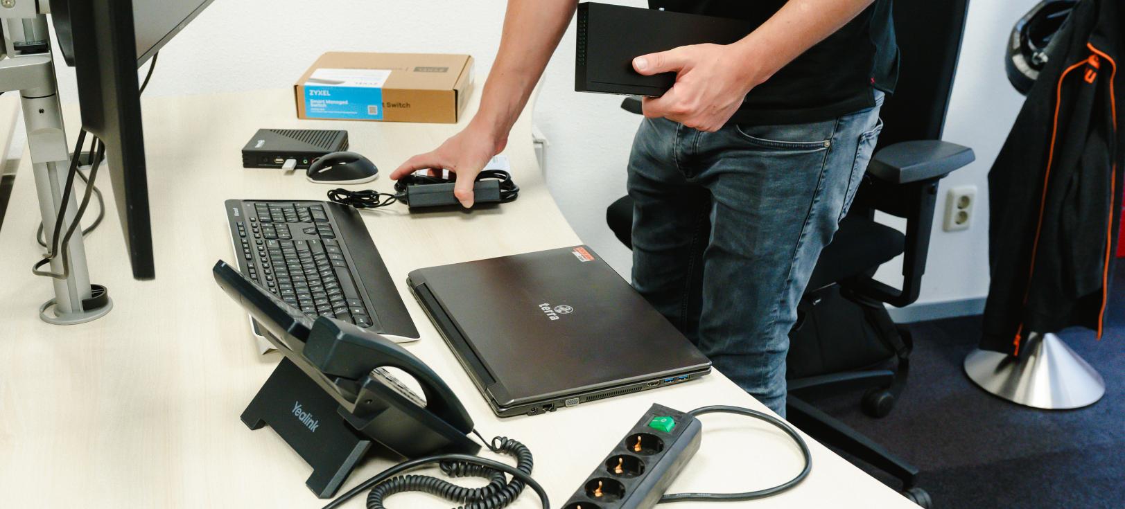 PWA medewerker die werkplekhardware instelt met laptop, toetsenbord, switch en telefonie op een bureau.