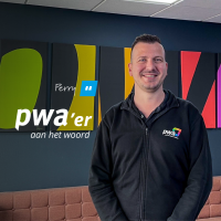 Perry, PWA’er aan het woord, lachend voor een kleurrijke wand in het PWA-kantoor.