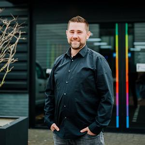 Man in zwarte blouse staat lachend voor de ingang van het PWA-pand, met het kleurrijke PWA-logo duidelijk zichtbaar boven de deur.