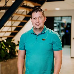 Man in turquoise poloshirt staat in de centrale hal van het PWA-kantoor, met een moderne trap en groene planten op de achtergrond.