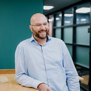 Man met bril en baard leunt lachend tegen een houten tafel in een moderne kantoorhal met groene wand en glazen scheidingswanden.