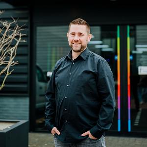 Man in zwarte blouse staat lachend voor de ingang van het PWA-pand, met het kleurrijke PWA-logo duidelijk zichtbaar boven de deur.