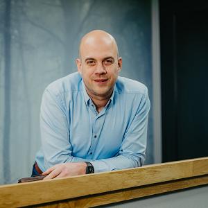 Man leunend op houten balustrade in modern kantoor, gekleed in lichtblauwe blouse, met rustige achtergrond van wanddecoratie met bosprint.