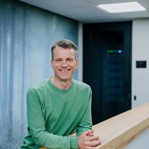 Man leunend op houten balustrade in modern kantoor, gekleed in lichtblauwe blouse, met rustige achtergrond van wanddecoratie met bosprint.