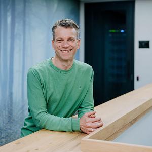 Man leunend op houten balustrade in modern kantoor, gekleed in lichtblauwe blouse, met rustige achtergrond van wanddecoratie met bosprint.