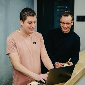 Twee collega’s die samenwerken achter een laptop aan een houten balustrade, met een serverruimte op de achtergrond.