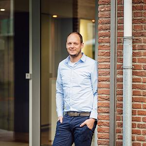 Man in lichtblauw overhemd staat ontspannen bij een kantoorgebouw met een bakstenen muur.