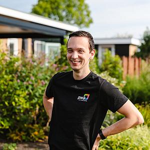 Lachende man in PWA-shirt poseert buiten voor een tuin met bloemen en struiken op een zonnige dag.