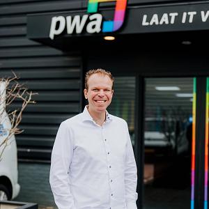 Medewerker van PWA staat glimlachend voor de ingang van het kantoor in Noordwijkerhout met PWA-logo boven de deur.