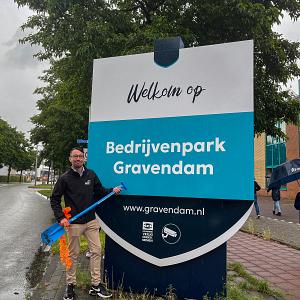 Man poseert met een grote symbolische sleutel voor een welkomstbord van Bedrijvenpark Gravendam, op een regenachtige dag met mensen op de achtergrond.