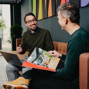 Twee mannen in gesprek op een bank in een kleurrijke kantooromgeving, met een open laptop en een brochure op schoot.