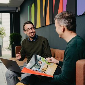 Twee mannen in gesprek op een bank in een kleurrijke kantooromgeving, met een open laptop en een brochure op schoot.
