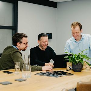 Drie mannen kijken samen naar een laptop aan een houten vergadertafel met een plant en glazen water in een moderne kantoorruimte.