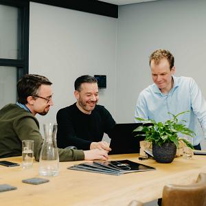 Drie mannen kijken samen naar een laptop aan een houten vergadertafel met een plant en glazen water in een moderne kantoorruimte.