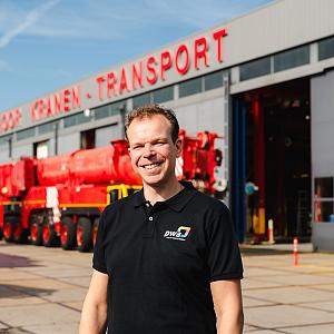 Man in zwarte polo met PWA-logo lacht voor een bedrijfspand met de tekst ‘Kranen - Transport’, met op de achtergrond een grote rode kraan en andere industriële voertuigen.