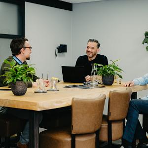Drie mannen zitten in gesprek aan een houten vergadertafel in een modern kantoor met planten en glazen water op tafel.