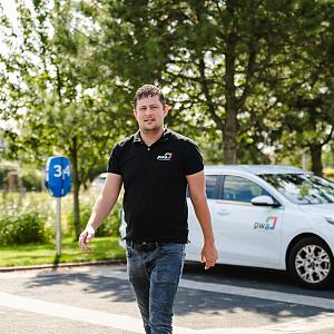 Man in PWA-bedrijfskleding loopt over een parkeerterrein met een PWA-auto en groene omgeving op de achtergrond.