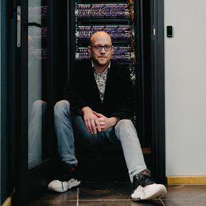 Man met bril zit ontspannen voor een open serverkast vol netjes weggewerkte paarse netwerkkabels in een moderne IT-ruimte.