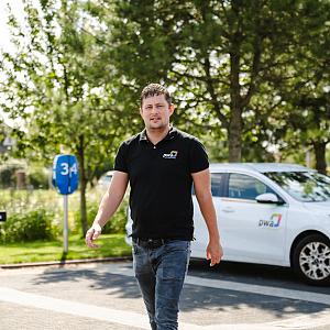 Man in PWA-bedrijfskleding loopt over een parkeerterrein met een PWA-auto en groene omgeving op de achtergrond.