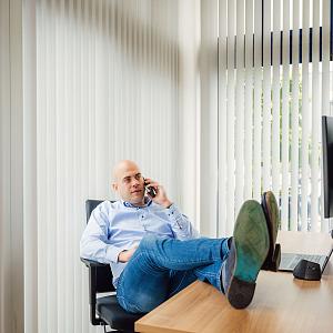 Man in PWA-shirt voert een telefoongesprek bij een raam met jaloezieën, waarop het logo van Novinco zichtbaar is.