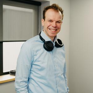 Man in lichtblauw overhemd leunend op een trapleuning, kijkend met een glimlach in een moderne kantooromgeving.