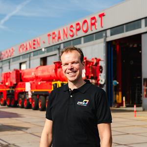 Lachende man in PWA-shirt staat voor een werkplaats van een kraan- en transportbedrijf met opvallende rode en blauwe voertuigen op de achtergrond.