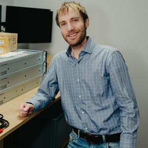 Man in een grijs overhemd, staand naast een bureau in een kantooromgeving.