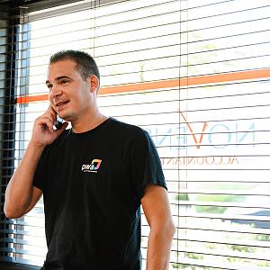 Man in PWA-shirt belt glimlachend bij het raam van een kantoor met jaloezieën en het logo van Novinco op de ruit.