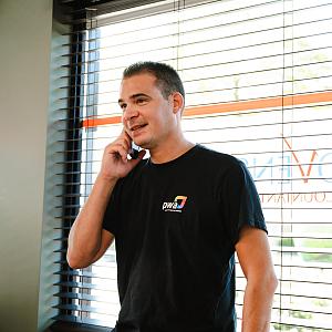 Man in PWA-shirt belt glimlachend bij het raam van een kantoor met jaloezieën en het logo van Novinco op de ruit.