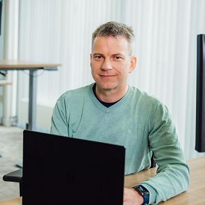 Lachende man in PWA-shirt staat voor een werkplaats van een kraan- en transportbedrijf met opvallende rode en blauwe voertuigen op de achtergrond.