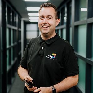 Man in PWA-bedrijfskleding staat lachend in een kantoorhal met glazen wanden en houdt een smartphone in zijn handen.