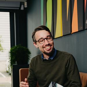 Lachende man in gesprek met een collega op een bank, met een brochure in de hand en kleurrijke kunst aan de muur op de achtergrond.