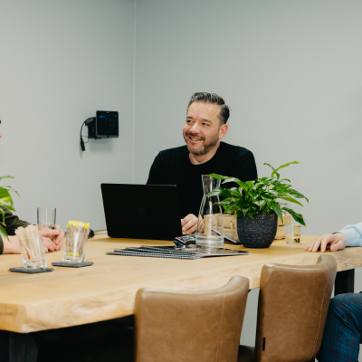 Drie mannen zitten aan een houten vergadertafel met een laptop, notitieboeken en glazen water, en hebben een informeel overleg in een moderne kantoorruimte.